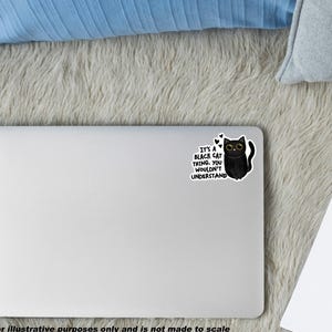 Op de afbeelding: Een zilveren laptop met een sticker van een zwarte kat met grote ogen en de tekst: It's a black cat thing. You wouldn't understand. De sticker bevat kleine hartjes boven de kat, die in een cartoonstijl is afgebeeld op de laptop.