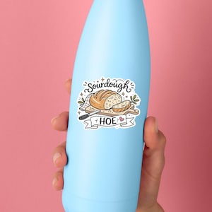 May include: A light blue water bottle featuring a sticker with an illustration of a loaf of sourdough bread on a wooden cutting board, a knife, and the text SOURDOUGH HOE written in black script and block lettering with a small pink heart icon.