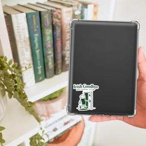 Puede incluir: Una tableta negra con una funda transparente, con una pegatina que dice "Irish Goodbye Anti Social Club". La pegatina representa una jarra de cerveza verde caminando por una puerta. Libros y una planta son visibles en el fondo.