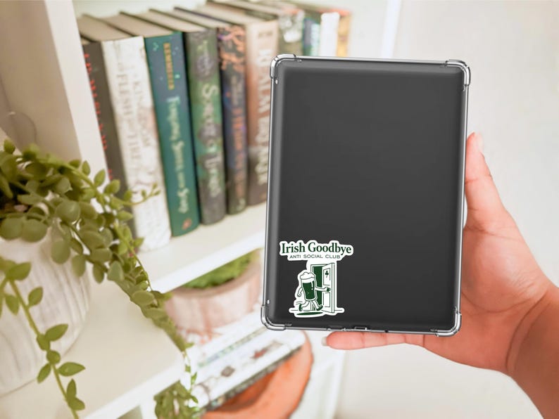 Puede incluir: Una tableta negra con una funda transparente, con una pegatina que dice "Irish Goodbye Anti Social Club". La pegatina representa una jarra de cerveza verde caminando por una puerta. Libros y una planta son visibles en el fondo.