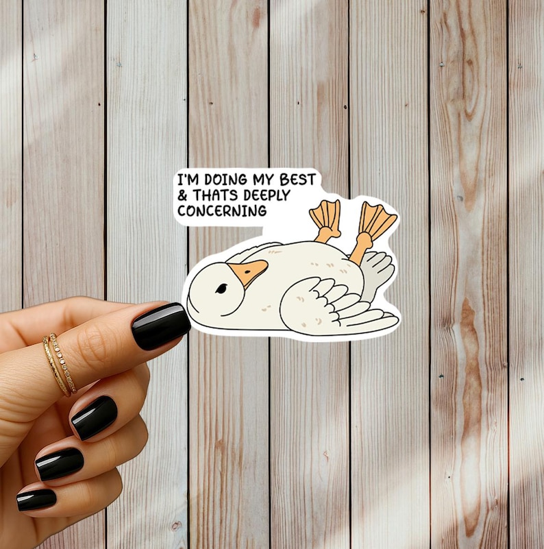 May include: A white sticker featuring a cartoon goose lying on its back with orange feet. The sticker has the text "I'M DOING MY BEST & THATS DEEPLY CONCERNING" in black font. The sticker is held by a hand with black nail polish.