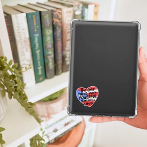 May include: A black tablet in a clear case, with a heart-shaped sticker that reads "The only thing more powerful than hate is love." The sticker has red, white, and blue accents. Bookshelves and plants are in the background.