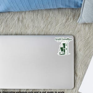Puede incluir: Un port&aacute;til blanco con una pegatina verde que dice "Irish Goodbye ANTI SOCIAL CLUB" con un dibujo animado de una jarra de cerveza saliendo por una puerta. El port&aacute;til est&aacute; sobre una superficie gris y esponjosa, con un coj&iacute;n azul y un bloc de notas.