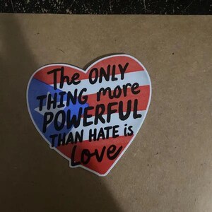 May include: Heart-shaped sticker featuring the Puerto Rican flag design and the words "The ONLY THING more POWERFUL THAN HATE is Love" in black and red. The sticker is on a brown surface.