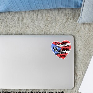 May include: A white laptop with a heart-shaped sticker. The sticker features the Puerto Rican flag design and the text "The ONLY THING more POWERFUL THAN HATE IS Love". The laptop is on a grey, fluffy surface.