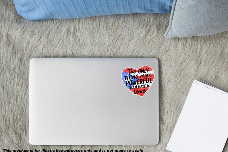 May include: A white laptop with a heart-shaped sticker. The sticker features the Puerto Rican flag design and the text "The ONLY THING more POWERFUL THAN HATE IS Love". The laptop is on a grey, fluffy surface.