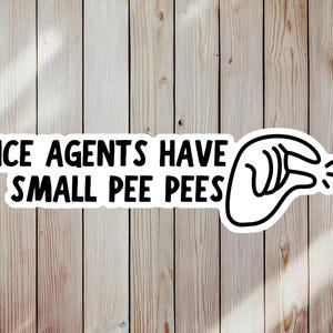 May include: A white sticker with black text that reads "ICE AGENTS HAVE SMALL PEE PEES" and a black and white graphic of a hand gesture. The sticker is on a light brown wooden surface.