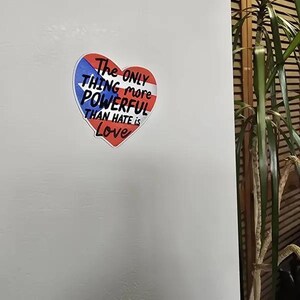 May include: A heart-shaped magnet with a red, white, and blue design. The magnet features the text "The ONLY THING more POWERFUL THAN HATE is Love" in black and white lettering. The magnet is attached to a white surface.
