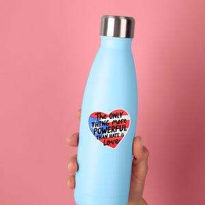 May include: A light blue insulated water bottle with a silver lid. A heart-shaped sticker on the bottle reads "The ONLY THING more POWERFUL THAN HATE is Love" in red, white, and blue.
