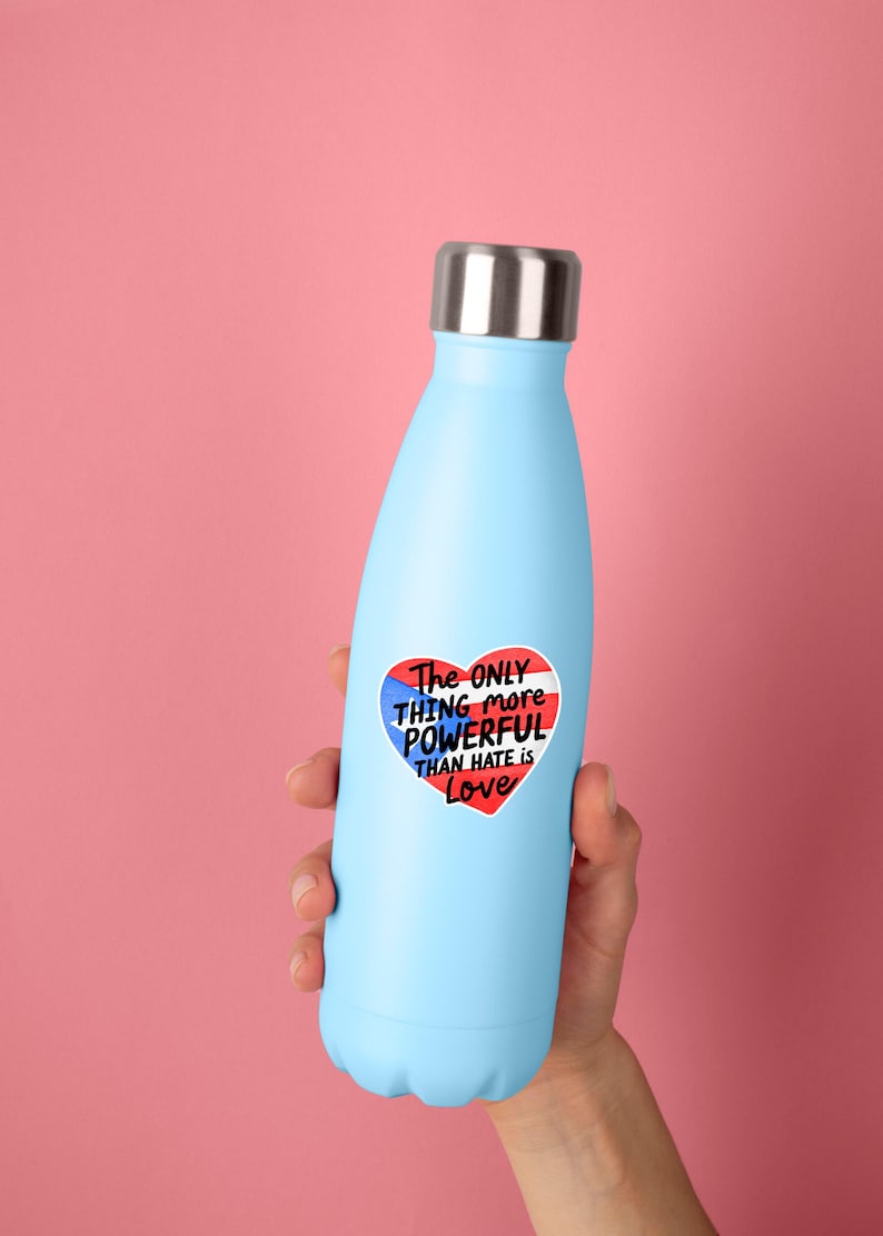 May include: A light blue insulated water bottle with a silver lid. A heart-shaped sticker on the bottle reads "The ONLY THING more POWERFUL THAN HATE is Love" in red, white, and blue.