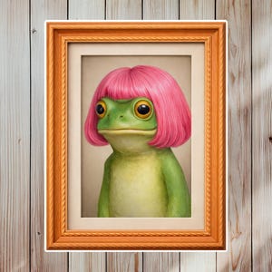 May include: A framed artwork featuring a green frog with large, expressive eyes and a pink wig. The frog is centered in a gold-colored frame, set against a neutral background. The artwork is displayed on a wooden surface.