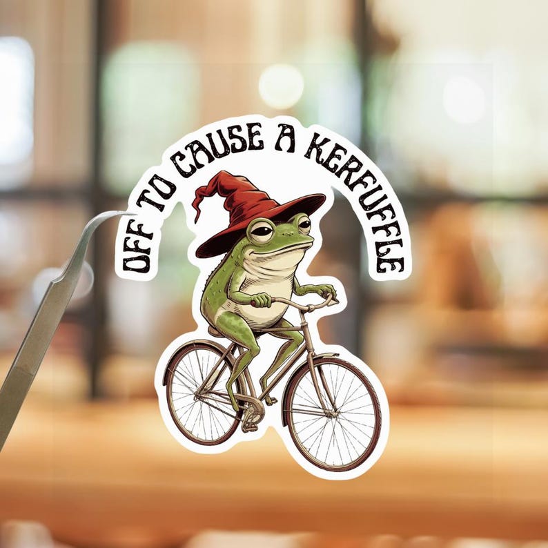 May include: A die-cut sticker featuring a green frog wearing a dark red pointed hat, riding a brown bicycle. The text "OFF TO CAUSE A KERFUFFLE" is arched above the frog in black lettering. The sticker is held by a pair of silver metal tweezers.