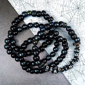 Könnte beinhalten: Vier schwarze Perlenarmbänder mit den Worten "DEAD INSIDE" auf einem der Armbänder. Die Armbänder liegen auf einem weißen Hintergrund mit einem Spinnennetz-Muster.