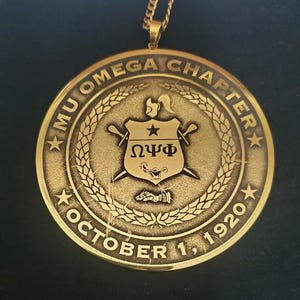 May include: Gold-toned pendant with the text "MU OMEGA CHAPTER OCTOBER 1, 1920". The pendant features a central emblem with a star, Greek letters, and crossed swords, surrounded by a laurel wreath design. The pendant hangs from a gold chain.