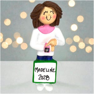 May include: A figurine of a woman wearing a white shirt with a pink collar and blue pants, pointing at a pink smartwatch. The figurine has brown hair and a smiling face. A green sign at the bottom reads "MADELINE 2020".