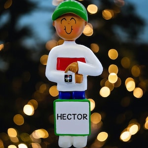 May include: A figurine of a person wearing a green cap, white sweater, blue pants, and white shoes. The figurine is holding a red book and pointing at a smartwatch. The name "HECTOR" is printed on a sign at the bottom.
