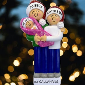 May include: A family Christmas ornament featuring a couple and a baby. The parents wear Santa hats with names on them. The baby is wrapped in a pink blanket. The family is wearing blue pants and white shirts. The base of the ornament reads "The CALLAHANS."