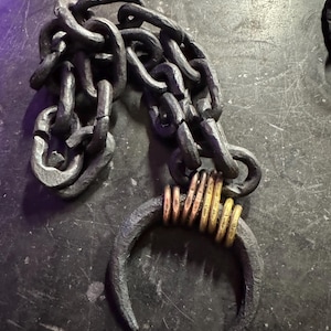 Może przedstawiać: Czarny metalowy łańcuch z dużym, zakrzywionym hakiem na końcu. Hak jest owinięty kilkoma mniejszymi, złotymi pierścieniami.
