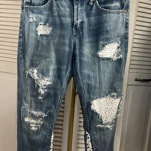 May include: Blue denim jeans with distressed details and floral fabric patches. The jeans have a classic five-pocket design and are hanging on a hanger. The distressed areas reveal patches of white fabric with a small floral pattern.