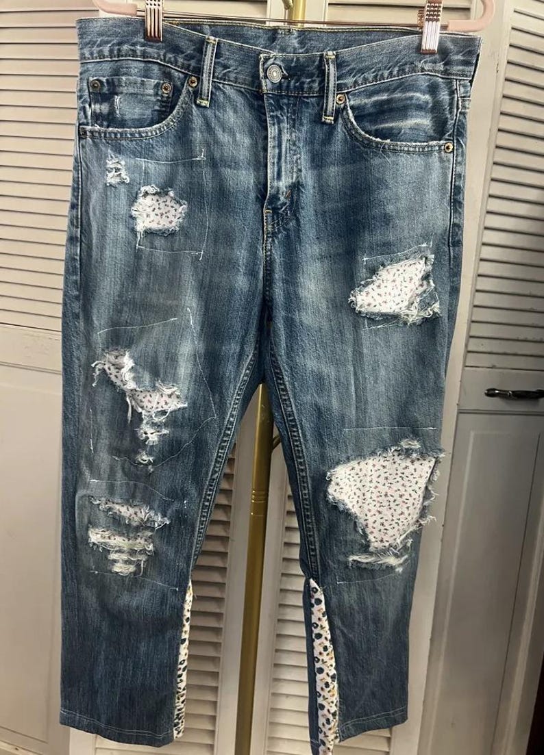 Puede incluir: Vaqueros azules con detalles desgastados y parches de tela floral. Los vaqueros tienen un dise&ntilde;o cl&aacute;sico de cinco bolsillos y est&aacute;n colgados de una percha. Las zonas desgastadas revelan parches de tela blanca con un peque&ntilde;o estampado floral.