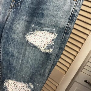 Puede incluir: Vaqueros de mezclilla azul descolorido con parches desgastados. Los parches est&aacute;n rellenos con una tela blanca con un peque&ntilde;o estampado floral en tonos rosas y rojos. Los vaqueros tienen un dise&ntilde;o cl&aacute;sico de cinco bolsillos.