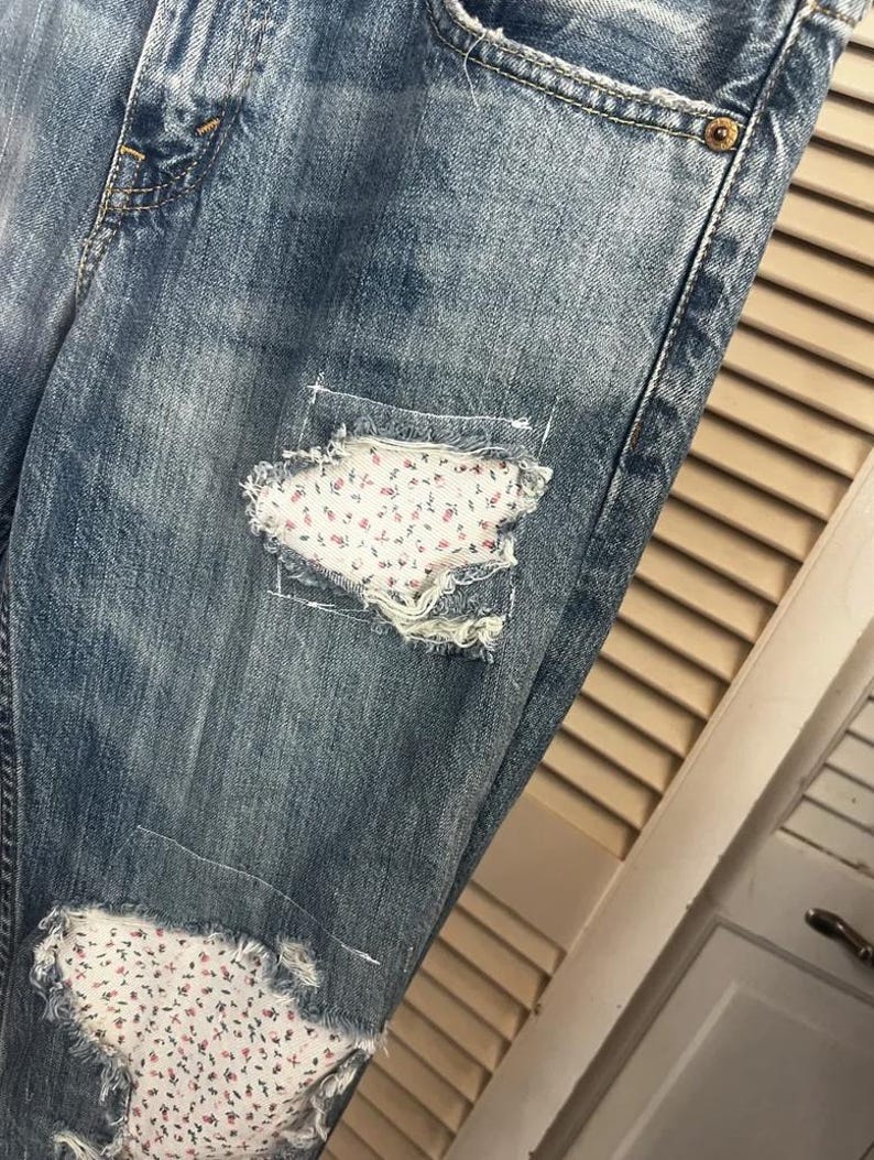 Puede incluir: Vaqueros de mezclilla azul descolorido con parches desgastados. Los parches est&aacute;n rellenos con una tela blanca con un peque&ntilde;o estampado floral en tonos rosas y rojos. Los vaqueros tienen un dise&ntilde;o cl&aacute;sico de cinco bolsillos.