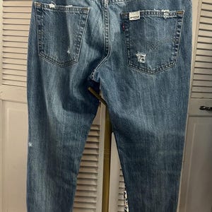 Puede incluir: Vaqueros de mezclilla azul descolorido con detalles desgastados, incluyendo rasgaduras y parches desgastados. Los bolsillos traseros muestran desgaste, y un parche de cuero Levi Strauss & Co. es visible. Los vaqueros tienen un dise&ntilde;o cl&aacute;sico de cinco bolsillos.