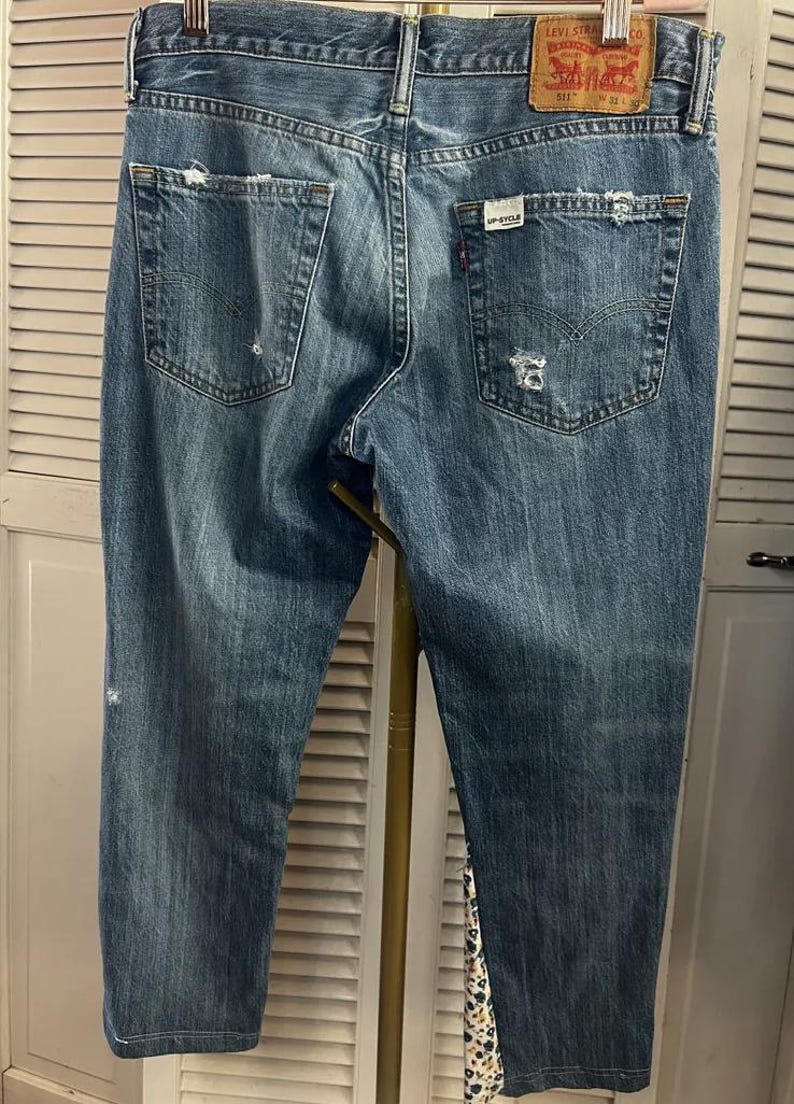 Puede incluir: Vaqueros de mezclilla azul descolorido con detalles desgastados, incluyendo rasgaduras y parches desgastados. Los bolsillos traseros muestran desgaste, y un parche de cuero Levi Strauss & Co. es visible. Los vaqueros tienen un dise&ntilde;o cl&aacute;sico de cinco bolsillos.