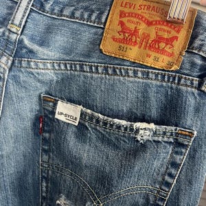 Puede incluir: Primer plano de unos vaqueros de mezclilla azules con un bolsillo desgastado y una etiqueta de cuero marr&oacute;n Levi Strauss. La etiqueta dice "511" y "W 31 L 30". Los vaqueros tienen el texto "UP-SYCLE" en el bolsillo.