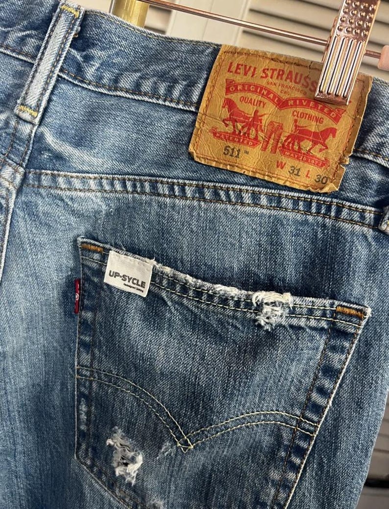 Puede incluir: Primer plano de unos vaqueros de mezclilla azules con un bolsillo desgastado y una etiqueta de cuero marr&oacute;n Levi Strauss. La etiqueta dice "511" y "W 31 L 30". Los vaqueros tienen el texto "UP-SYCLE" en el bolsillo.