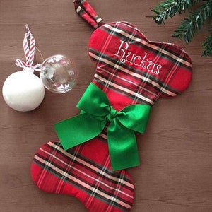 May include: A red plaid Christmas stocking with a green satin bow and the name "Ruckus" embroidered in white. Two Christmas ornaments hang to the left of the stocking. A festive holiday decoration.