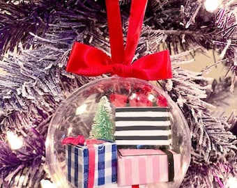 May include: A clear Christmas ornament with miniature gift boxes and a small green tree inside. The ornament is suspended by a red velvet ribbon and bow, set against a frosted purple Christmas tree.