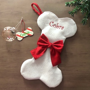 May include: A white felt dog bone-shaped Christmas stocking with a red bow and the name "Cobra" embroidered in red. A matching bone-shaped ornament and a clear glass ball ornament are also present.