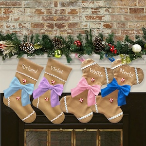 May include: Four gingerbread-shaped Christmas stockings with names embroidered on them: Richard, Violet, Waffle, and Mochi. Each stocking has a colorful bow and a candy button. They are hanging above a fireplace decorated with garland.