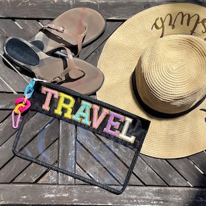 May include: A clear plastic zippered pouch with a black fabric flap. The flap has the word "TRAVEL" embroidered in colorful letters. The pouch is lying on a wooden surface with a straw hat and sandals.