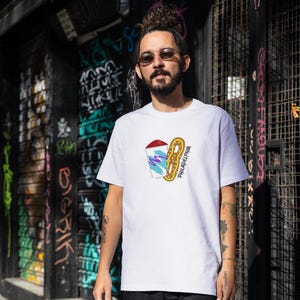 Op de afbeelding: Wit T-shirt met een grafisch ontwerp van een drankje, een pretzel en het woord "PHILADELPHIA". Het ontwerp gebruikt rode, paarse, gele en blauwe kleuren. Het T-shirt is een klassieke ronde hals.