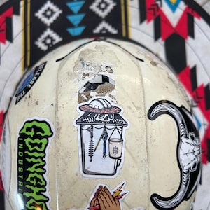May include: A well-worn, off-white hard hat adorned with various stickers. The stickers include a green, stylized text that reads "Ginchy Industries", a skull with a halo, praying hands with lightning bolts, and a crescent moon skull. The brim features a geometric pattern.