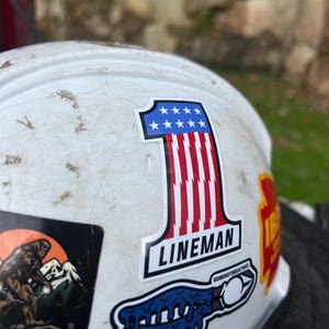 May include: A white hard hat with several stickers. The prominent sticker is a number one with red and white stripes and blue stars, with the word "LINEMAN" below. Other stickers include a Bigfoot graphic and a blue dripping design.