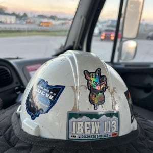 May include: A white hard hat with multiple stickers, including one that reads "IBEW 113 Colorado Springs." The hat is worn and shows signs of use. The background shows a vehicle interior and a blurred outdoor scene.
