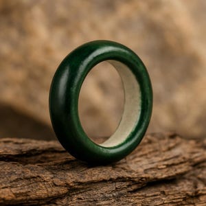 May include: A dark green ring with a smooth, polished exterior. The ring is set against a textured, brown wooden surface. The interior of the ring is a lighter, off-white color, creating a contrast with the dark green.