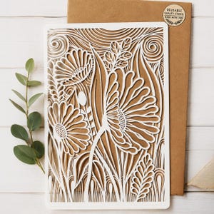 May include: A white rectangular stencil featuring a laser-cut floral design with large daisy-like flowers, swirling lines, and foliage patterns. The stencil is placed on a brown paper envelope with a circular sticker that reads Reusable Quality Stencil.