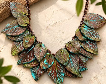May include: A statement necklace featuring multiple leaf-shaped pendants attached to a copper-toned chain. Each leaf has a textured surface with a metallic brown finish and vibrant turquoise blue accents highlighting the intricate vein patterns.