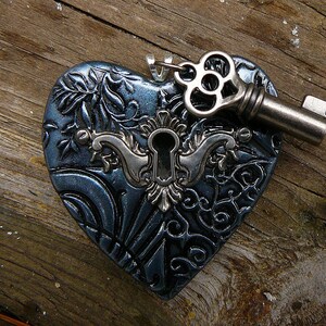 May include: A dark blue heart-shaped pendant with an intricate design and a keyhole. A silver key with a decorative head is attached to the pendant.