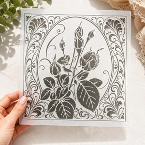 Puede incluir: Una carpeta de relieve cuadrada de plástico translúcido con un diseño impreso en gris oscuro de capullos de rosa y hojas encerrados en un marco ovalado. El borde está decorado con intrincados patrones de volutas simétricas en las esquinas.