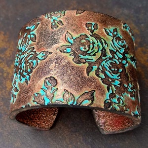 Pode incluir: Uma pulseira larga com um padrão floral. A pulseira é de cor cobre com detalhes turquesa, e o desenho floral é em relevo. A pulseira tem um acabamento texturizado e é feita de um material resistente.