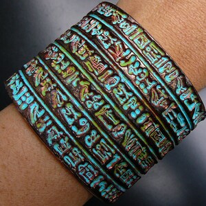May include: A wide copper bracelet with multiple bands, each featuring a raised design of ancient Egyptian hieroglyphs. The bands are painted in shades of turquoise, green, and brown.