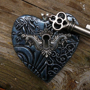 May include: A dark blue heart-shaped pendant with an intricate design and a silver keyhole. The keyhole is surrounded by a silver filigree design. A silver key with a decorative head is attached to the pendant.