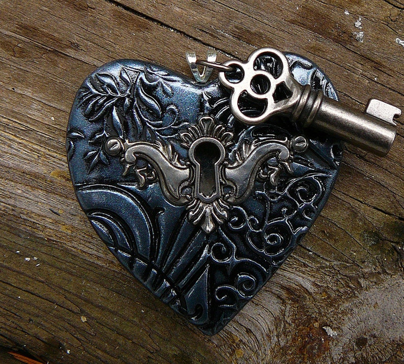 May include: A dark blue heart-shaped pendant with an intricate design and a silver keyhole. The keyhole is surrounded by a silver filigree design. A silver key with a decorative head is attached to the pendant.