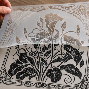 May include: A translucent plastic stencil sheet featuring a detailed floral design with calla lilies and ornate border patterns. The stencil is partially lifted to reveal a crisp black ink print of the same botanical illustration on paper.