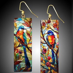 May include: A pair of stained glass earrings featuring colorful birds perched on branches with red flowers. The birds have blue, orange, and yellow bodies with long blue tails.